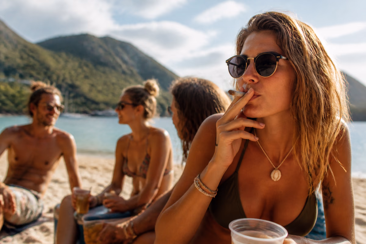 Vrouw met zonnebril rookt een joint op een tropisch strand, met vrienden met drankjes op de achtergrond