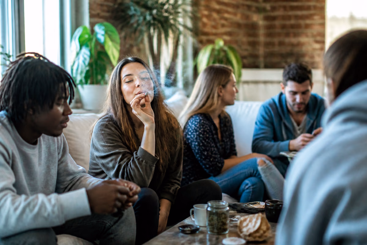 Jongvolwassenen op een bank, een vrouw rookt een joint, op tafel een pot met cannabistoppen