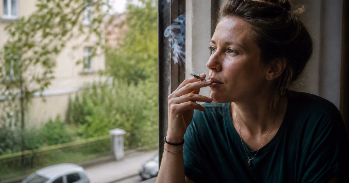 Vrouw rookt een joint bij het raam en kijkt uit op een groene straat